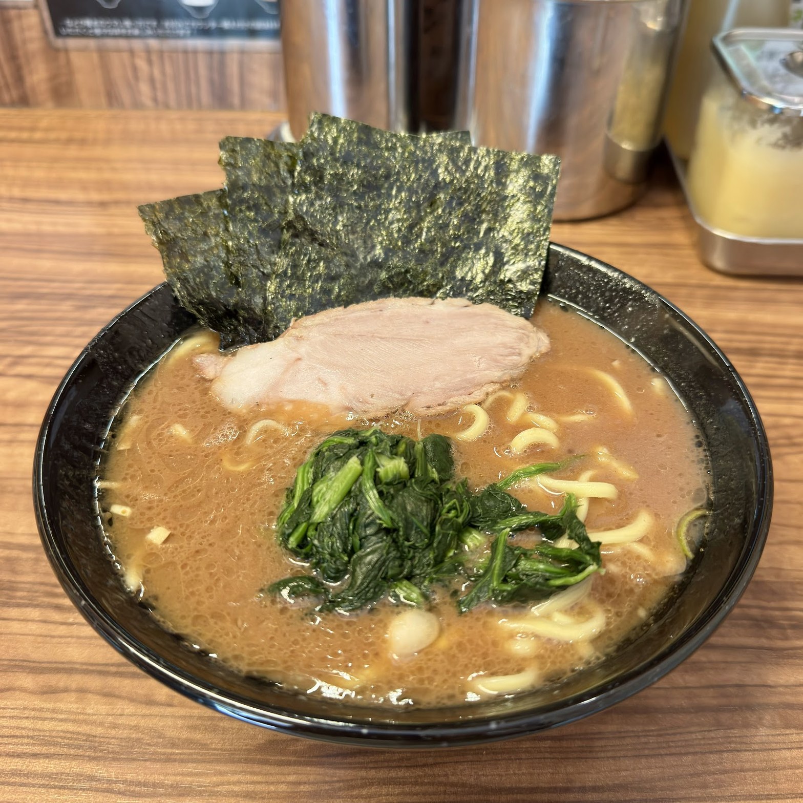 黒い丼に盛られた家系ラーメン、海苔3枚・チャーシュー・ほうれん草・味玉が乗り、茶色い豚骨醤油スープが表面に油膜を張っている